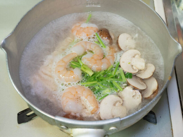 鍋にお湯を沸かして、インスタント麺、むきエビ、マッシュルーム、パクチーの茎を加えた写真